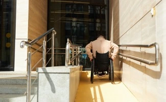 Una persona en silla de ruedas asciende por una rampa amplia para acceder al edificio de su vivienda.