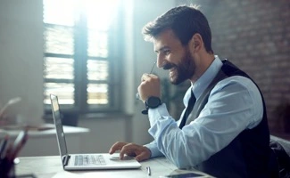 Hombre sonriente trabajando en un portátil en una oficina luminosa, apoyando la mano en el rostro mientras se concentra en la pantalla.