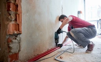 Trabajador realizando obras en el interior de una vivienda, perforando una pared para instalar conducciones, en un entorno de reforma o mantenimiento.