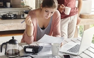 Mujer sentada en una mesa revisando facturas y documentos financieros junto a una computadora portátil, con una cafetera y una taza de café, mientras otra persona de pie sostiene una taza al fondo en un ambiente doméstico.