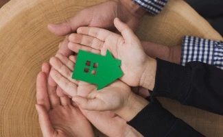 Manos de un adulto y un niño sosteniendo una figura de casa verde, simbolizando protección del hogar, seguridad o cobertura como un seguro de vivienda.