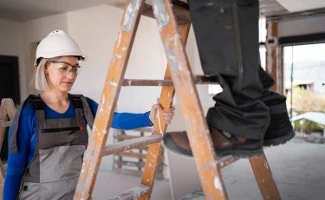 Trabajadores realizando una obra en una vivienda o zona común, utilizando una escalera y equipados con casco y ropa de seguridad.