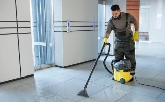 Trabajador de limpieza utilizando una aspiradora industrial para limpiar el suelo de un edificio moderno con ascensor y pasillos amplios.